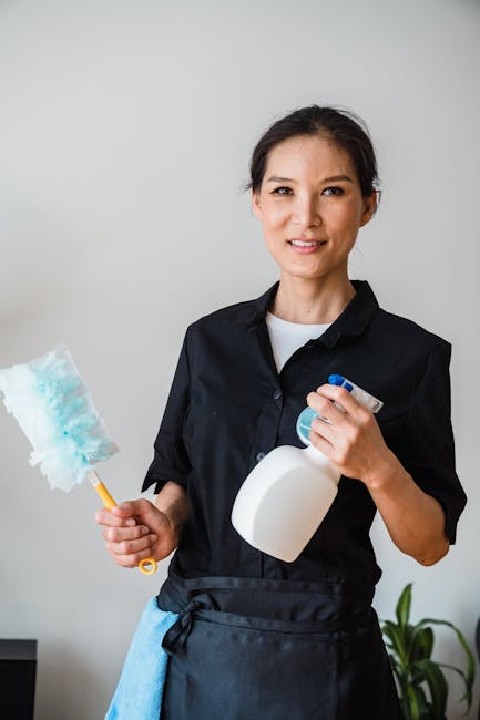 A professional cleaner from Carpet Cleaners Belgravia stands indoors in a well-lit room with neutral-colored walls. She holds a cleaning spray bottle in one hand and a blue and white scrubbing brush i