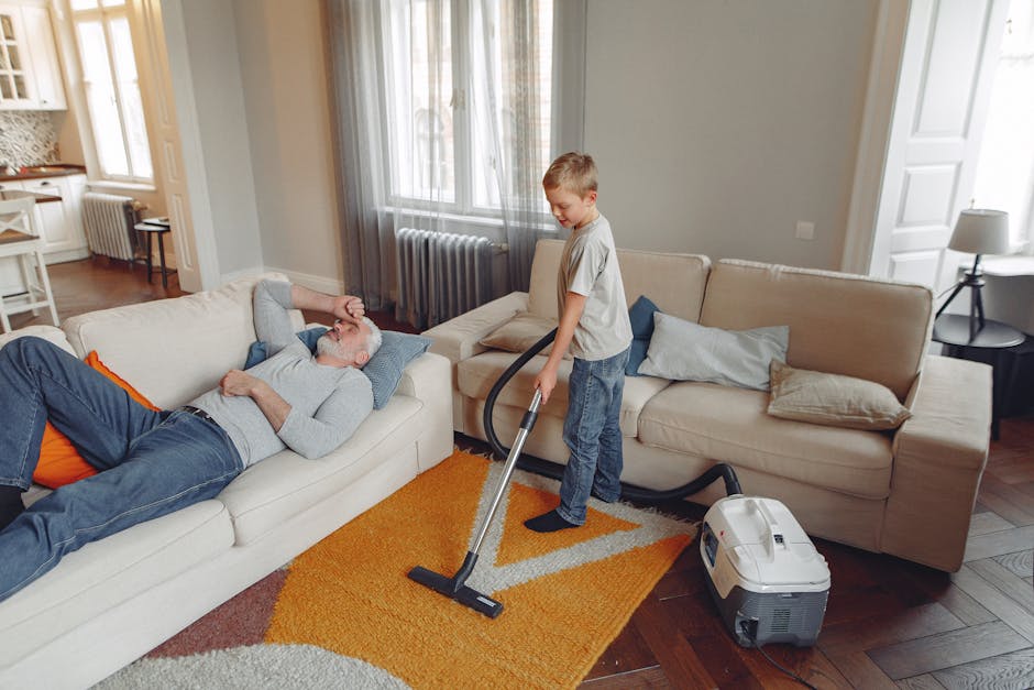 Close-up of a red and black vacuum cleaner with a transparent dust container filled with dust, connected to a flexible hose and a floor attachment. The vacuum is positioned on a beige carpet, with a wooden skirting board visible along the wall. The scene takes place in a well-lit room, emphasizing the process of surface cleaning or deep cleaning in a domestic setting. The equipment appears modern and well-maintained, with the vacuum’s hose and floor tool in contact with the carpet when functioning, highlighting hygienic surface maintenance as part of professional cleaning services offered by Carpet Cleaners Belgravia for period homes in Eaton Square.