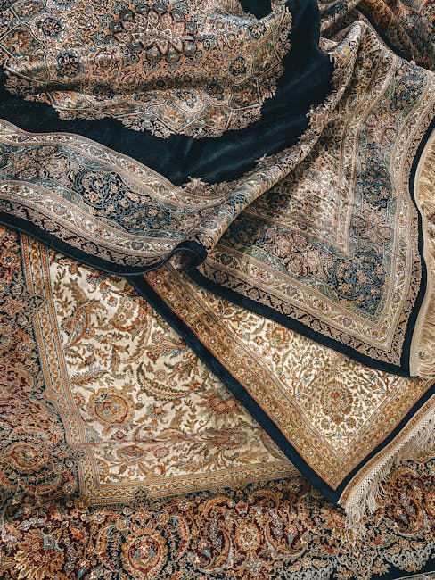 A close-up view of multiple intricately patterned Oriental rugs with detailed floral and geometric designs, layered over each other, showcasing their rich colors including beige, gold, navy blue, and 