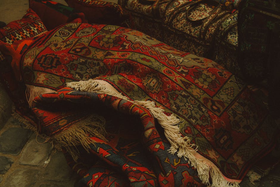 A close-up view of multiple intricately patterned Oriental rugs with detailed floral and geometric designs, layered over each other, showcasing their rich colors including beige, gold, navy blue, and 